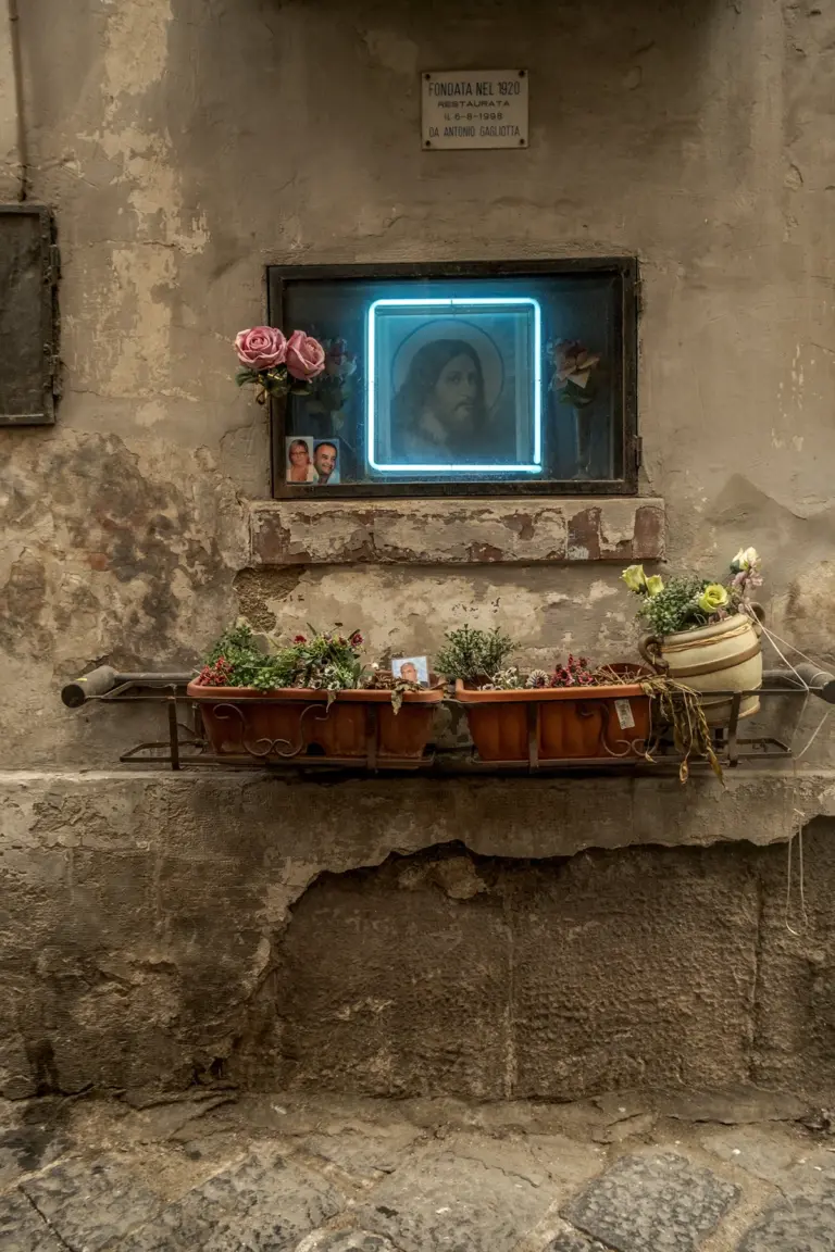 Altar callejero en Nápoles con icono religioso enmarcado en neón azul, flores y fotografías de difuntos sobre una repisa en una fachada deteriorada