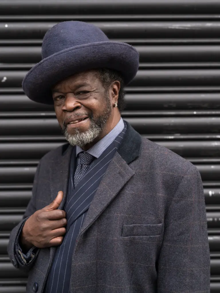 Ridley Road smells of spices and fire Hombre con sombrero y abrigo de tweed posando frente a una persiana metálica, serie Ridley Road smells of spices and fire